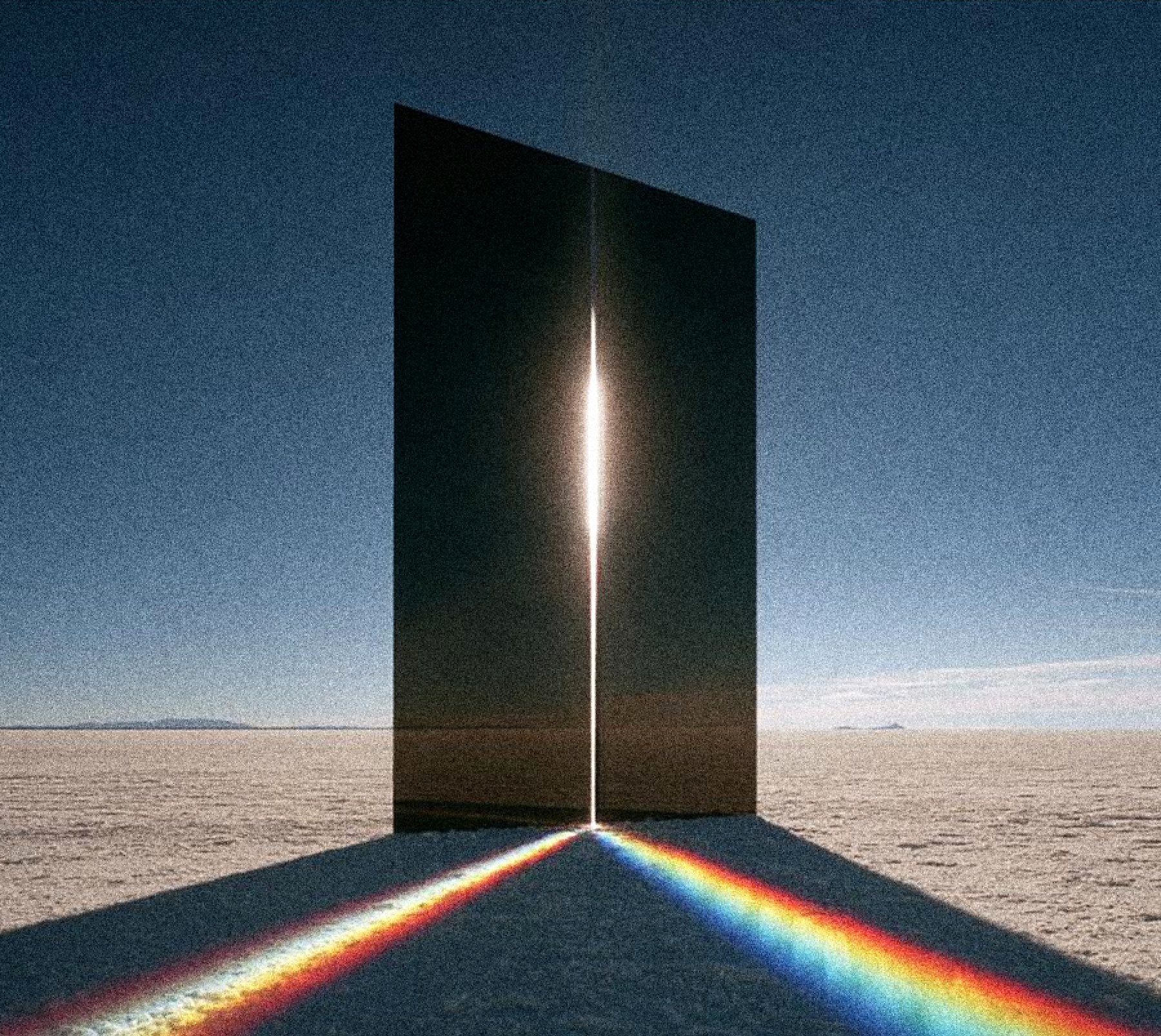 A tall dark glass monolith on a white salt desert, a slit of white light and a spectrum fanning across the salt